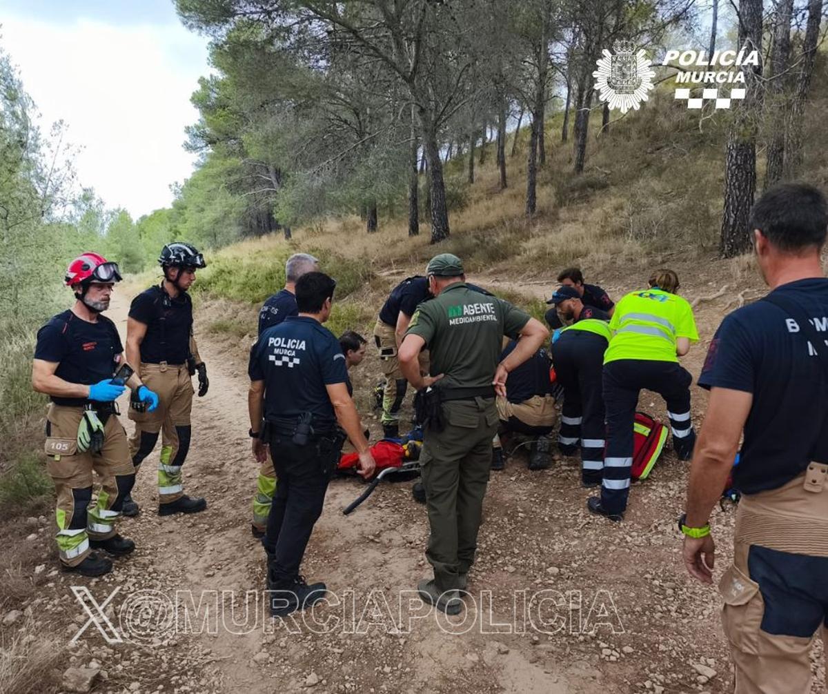 Varios policías locales y bomberos ayudan a subir al ciclista herido a una camilla.