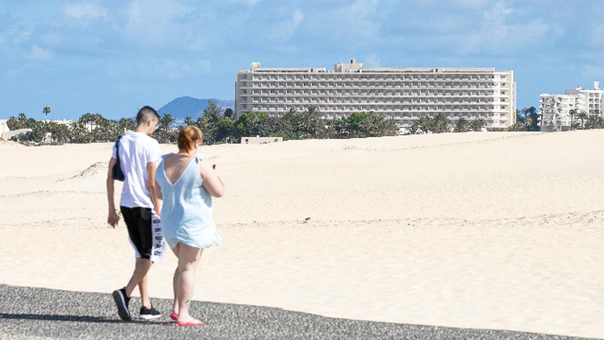 Dos turistas pasean hacia las Dunas de Corralejo, con el Hotel OlivaBeach al fondo.