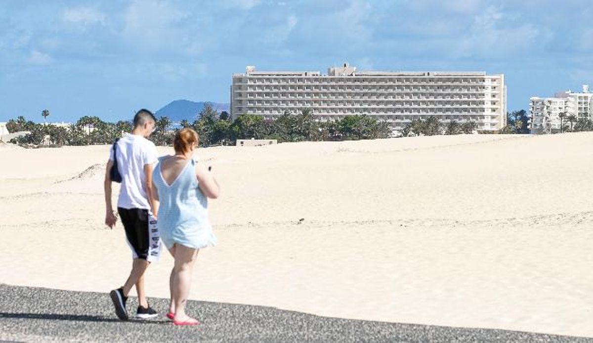 Two tourists walk towards the Corralejo Dunes, with the OlivaBeach Hotel in the background.