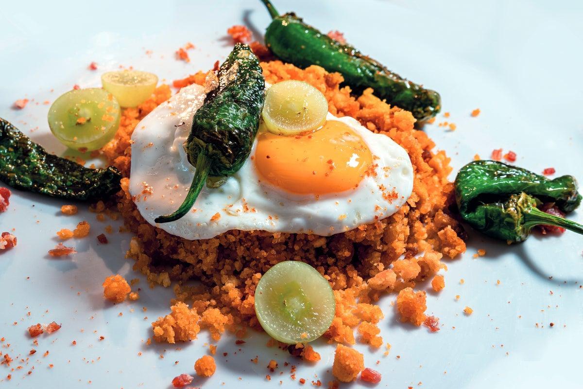 Migas rondeñas: Desmigar el pan duro y dejarlo toda la noche con un poco de caldo de puchero, pimentón, sal y pimienta. Refreír con chorizo y panceta y servir con los huevos camperos, uvas, pimientos de Padrón y decorado con una arena de jamón ibérico frito.
