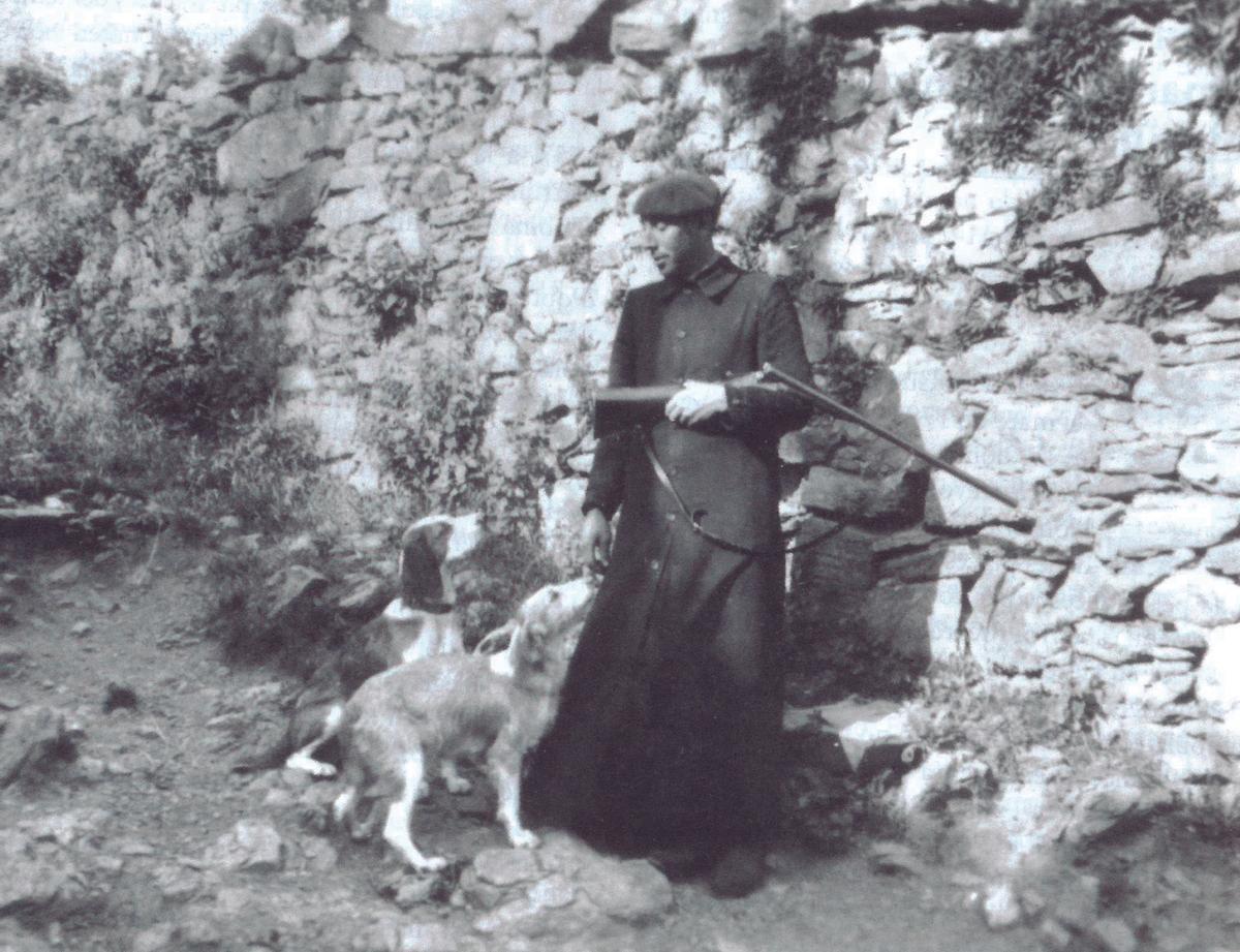 Mossèn Lluís Suriñac de Rocabruna amb sotana, boina, escopeta i els gossos, en un dia de cacera, als anys 50.