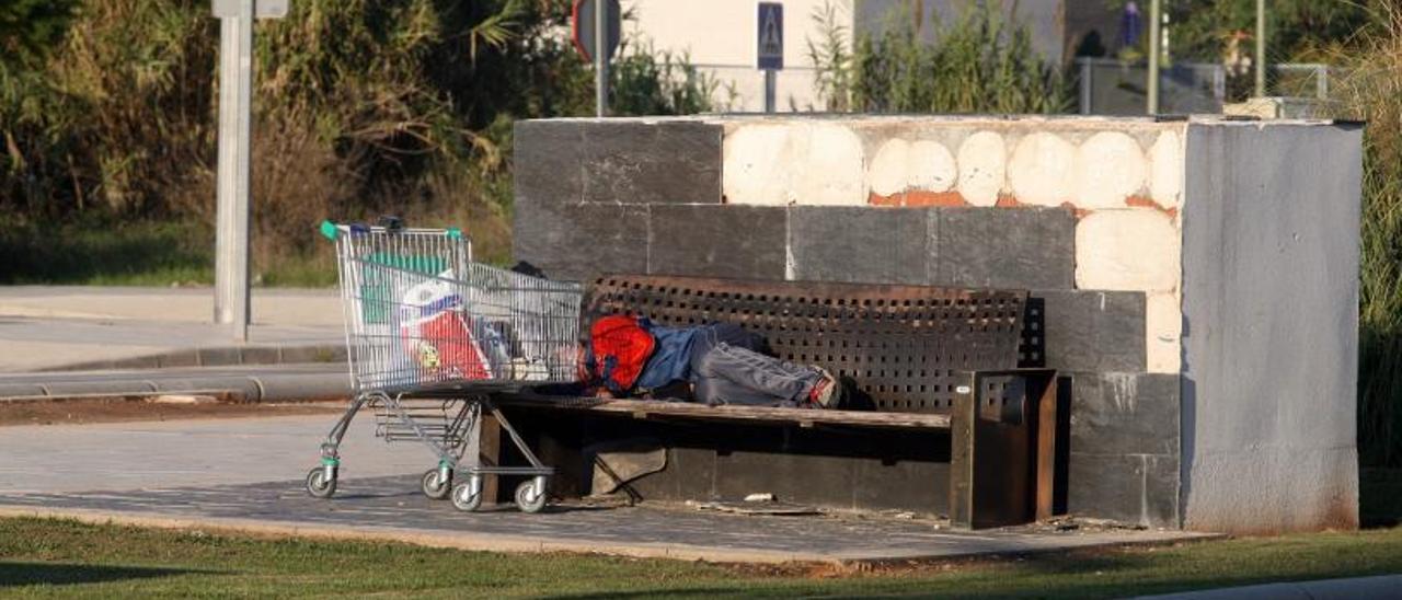 Un joven, acostado en un banco de Alzira junto a sus pertenencias, que guarda en un carro de la compra de un supermercado, en una imagen de archivo. | VICENT M. PASTOR