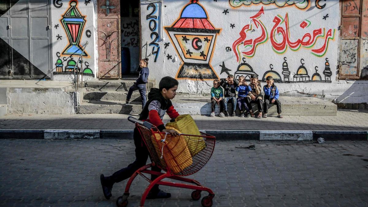 Un niño palestino empuja un carro con bidones de agua frente a un muro con dibujos temáticos del Ramadán en la ciudad de Gaza, este viernes.