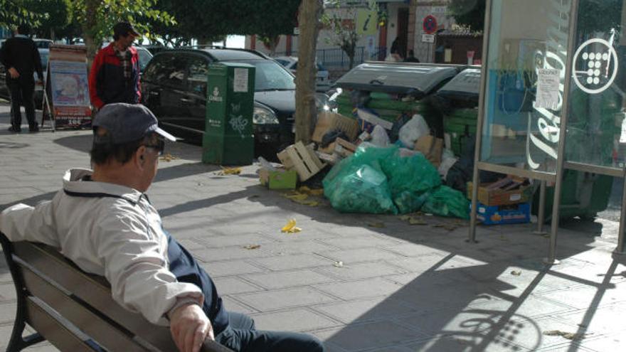 Un vecino toma el sol en un banco de una calle de Rincón ante unos contenedores atestados de bolsas de basura.