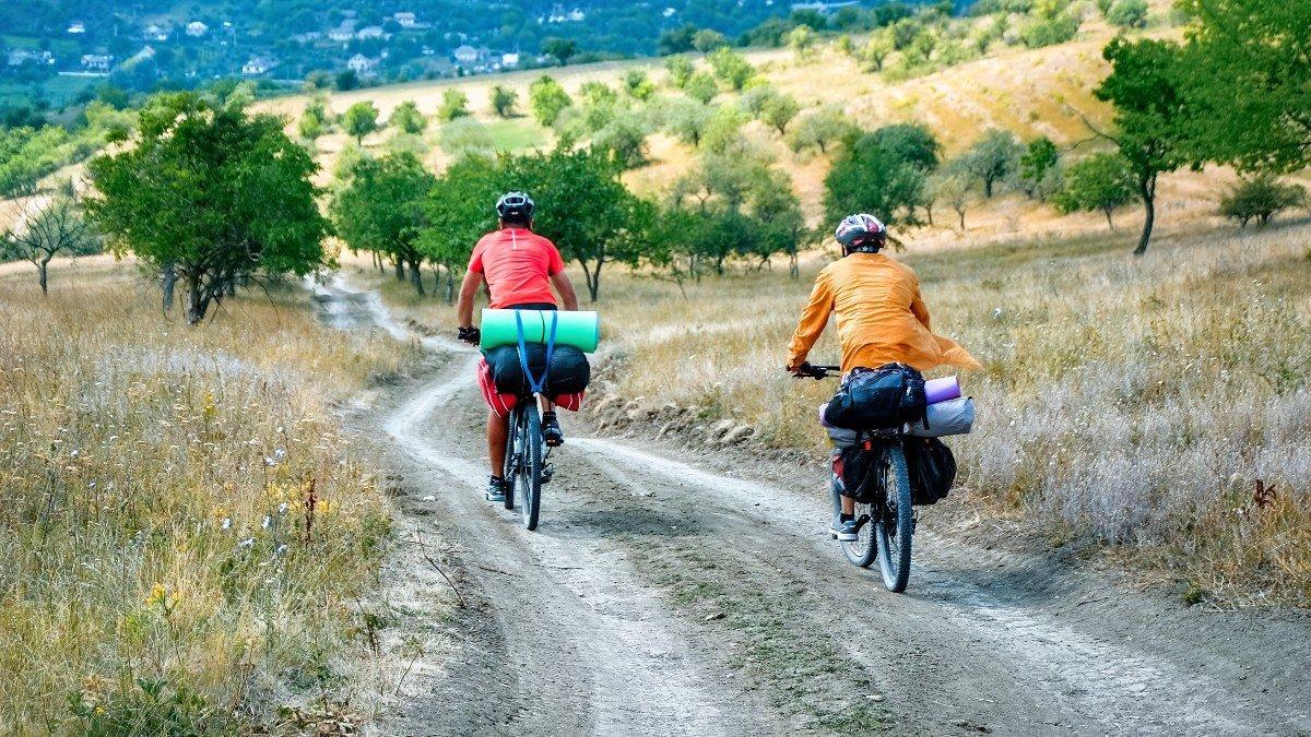 two-cyclists-in-helmets-with-bicycles-full-of-traveler-s-stuff-moving-on-the-country-road-through-rare-green-trees