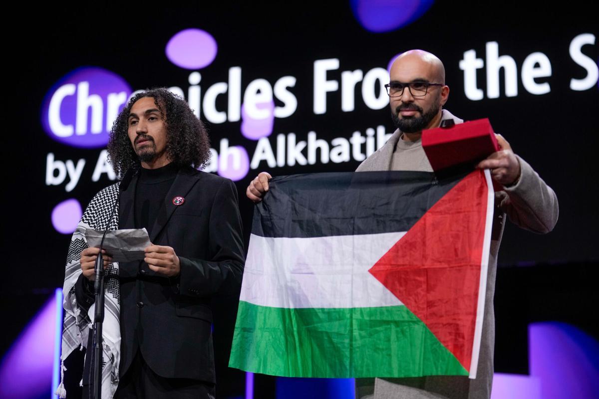 El director Abdallah Al-Khatib, a la izquierda, se dirige al público tras recibir el premio GWFF a la mejor ópera prima por "Chronicles from the Siege", mientras Taqiyeddine Issaad, a la derecha, sostiene una bandera palestina durante la ceremonia de entrega de premios del Festival Internacional de Cine de Berlín