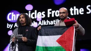 El director Abdallah Al-Khatib, a la izquierda, se dirige al público tras recibir el premio GWFF a la mejor ópera prima por Chronicles from the Siege, mientras Taqiyeddine Issaad, a la derecha, sostiene una bandera palestina durante la ceremonia de entrega de premios del Festival Internacional de Cine de Berlín