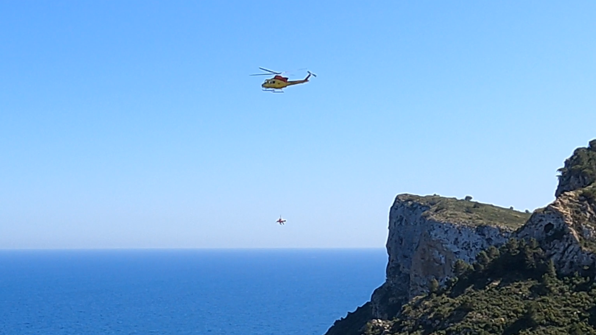 El helicóptero, con el herido en camilla y un bombero con él, sobrevuela la cala de Llebeig. | RICARDO REIG/LA SENDA TUERTA