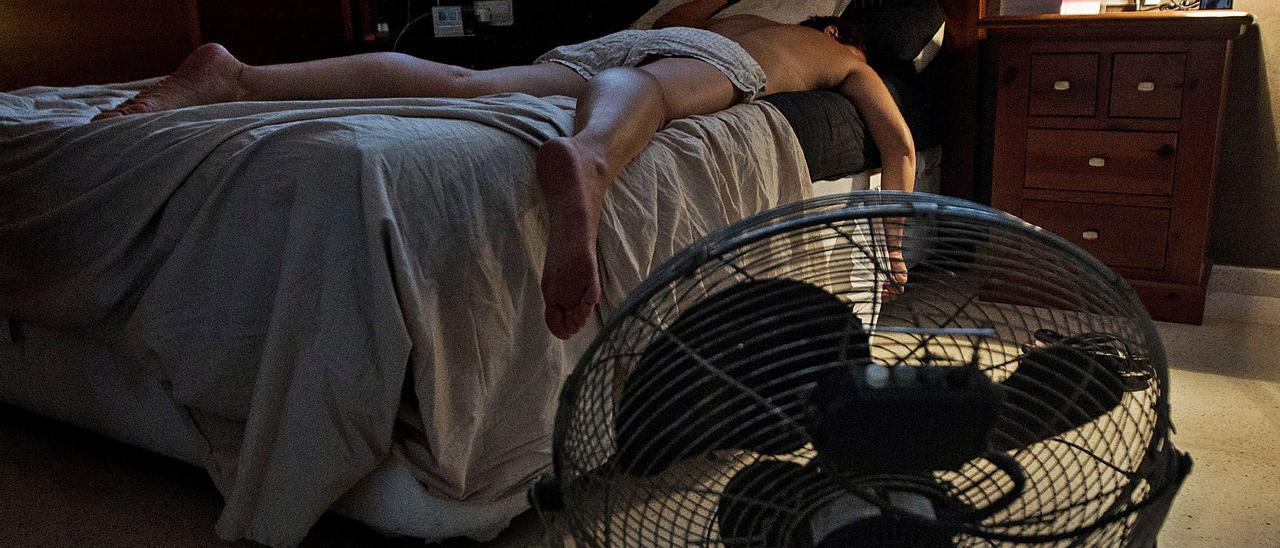 Un joven duerme echado sobre la cama junto a un ventilador para tratar de mitigar el calor nocturno de Alicante.