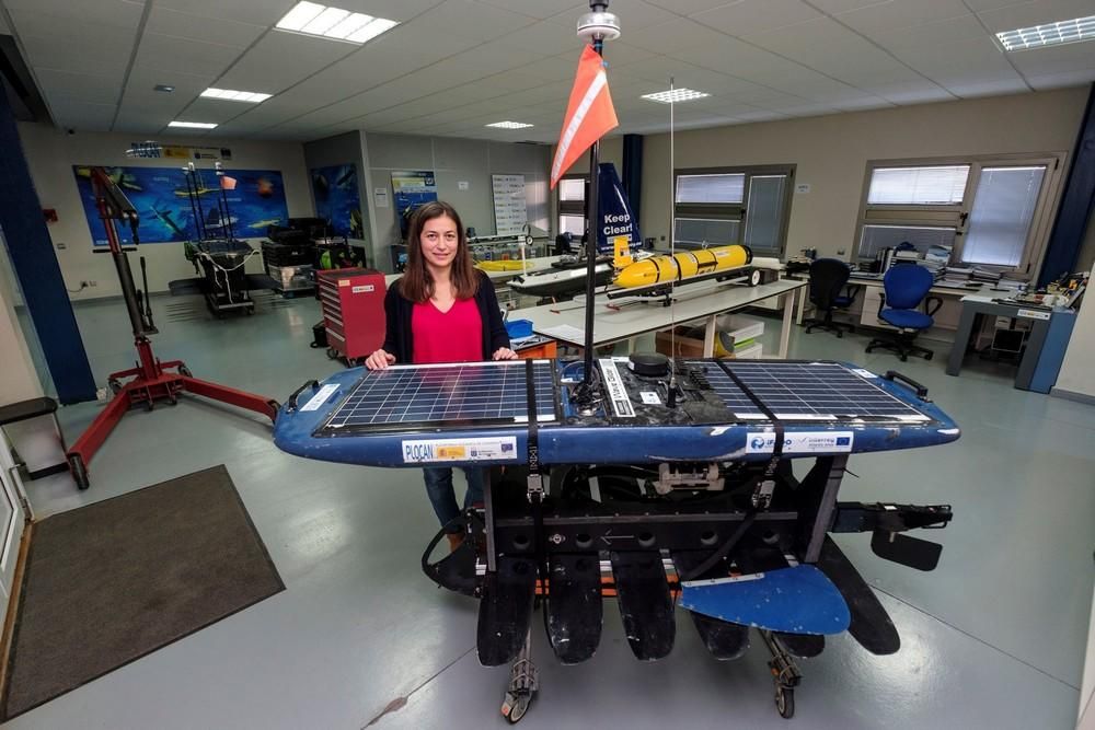 La ingeniera de Robótica de la Plataforma Oceánica de Canarias (Plocán), Tania Morales, muestra el "wave glider" (un robot que navega por la superficie impulsado por la energía de las olas) que ha recorrido en los dos últimos meses 2.000 kilómetros entre los archipiélagos de Azores, Madeira y Canarias recopilando datos sobre el océano, los cetáceos y el ruido del tráfico marítimo. Detrás de ella, un planeador submarino (en amarillo) y un navegador de superficie a vela (azul).