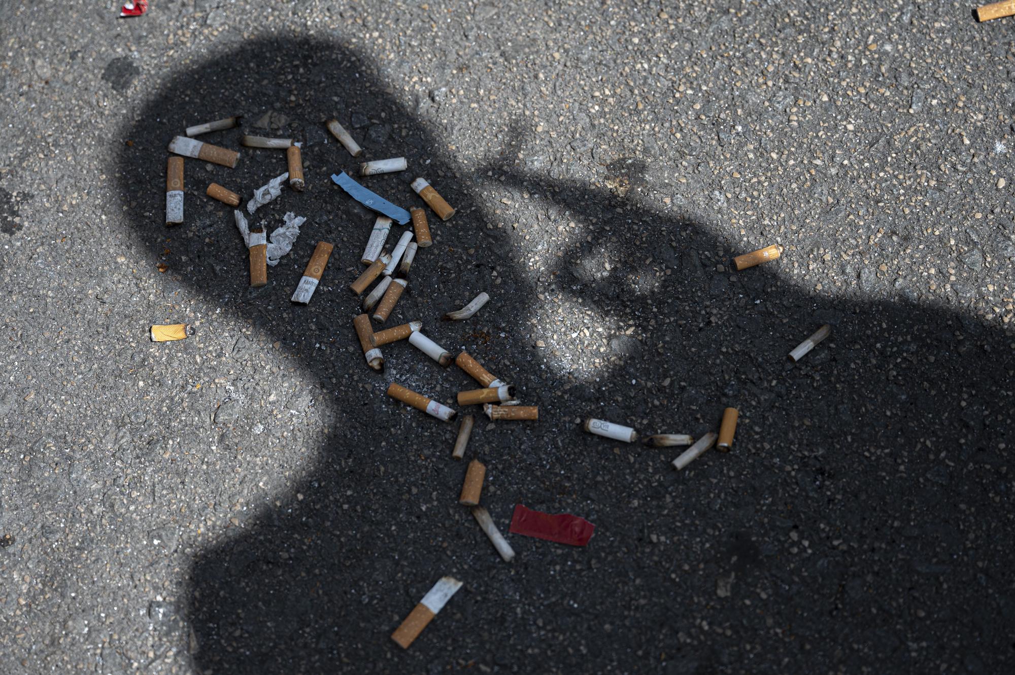 La sombra de un hombre fumando se refleja sobre un puñado de colillas tiradas sobre la acera.