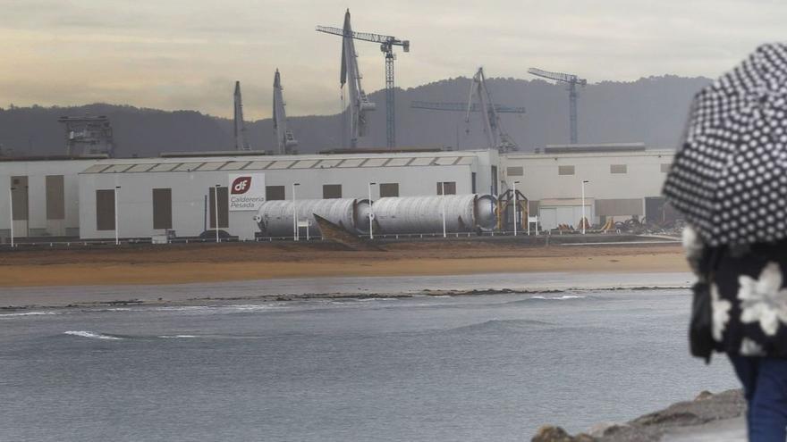 Una mujer pasea junto a la playa de Poniente de Gijón, con el Tallerón de Duro Felguera al fondo. | ÁNGEL GONZÁLEZ