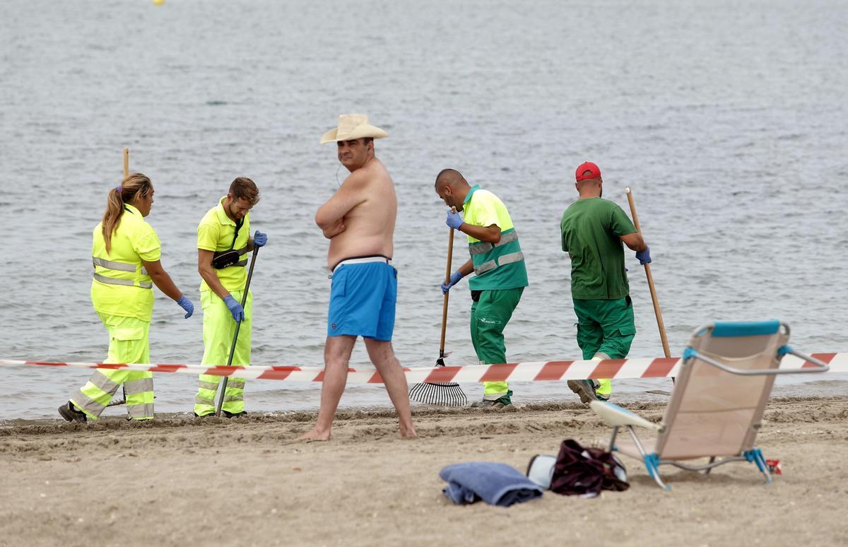 Turistas observan como operarios de La Línea retiran restos de aceite de la Playa de Poniente.