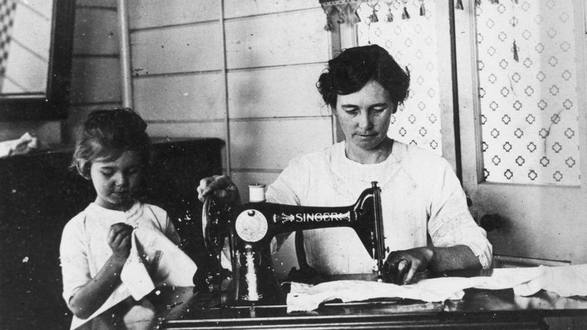 Una madre junto a su hija cosiendo con una máquina Singer en el pasado.