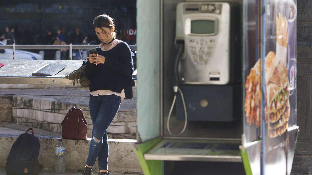 Una joven mira su teléfono móvil junto a una cabina telefónica.