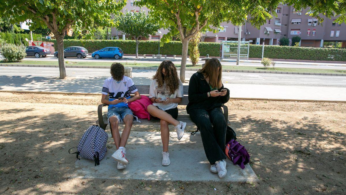 Tres jóvenes utilizan el móvil sentados en un banco.