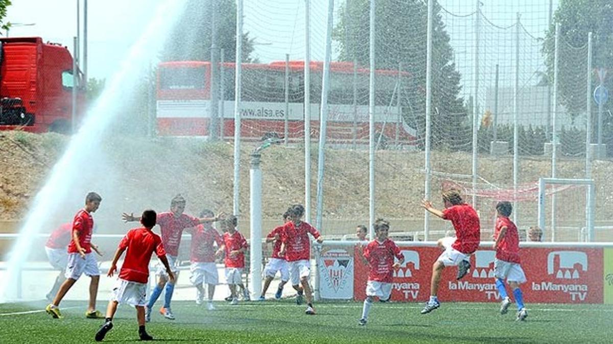Un grup de nens futbolistes de l’Atlètic Segre de Lleida juguen sota les regadores del camp.
