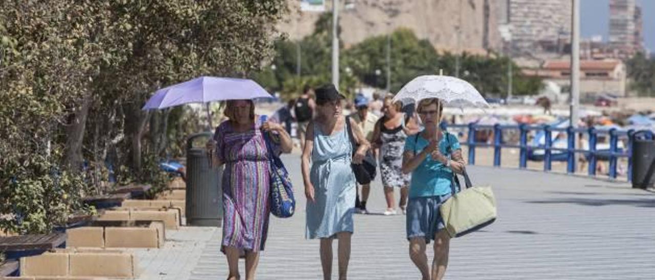 Tres mujeres, dos de ellas cubriéndose del intenso sol con sendos paraguas, pasean por la zona de la playa del Postiguet