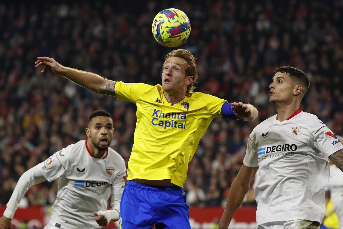 SEVILLA, 21/01/2023,- El centrocampista del Cádiz Álex Fernández (c) cabecea el balón ante los jugadores del Sevilla Youssef En-Nesyri (i) y Erik Lamela, durante el partido de la jornada 18 de LaLiga que estos dos equipos juegan hoy sábado en el estadio Sánchez Pizjuán. EFE/ Julio Muñoz