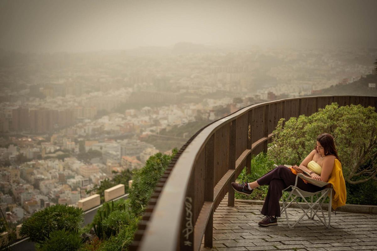Una mujer lee en un mirador desde el que se puede contemplar la espesa calima sobre Santa Cruz