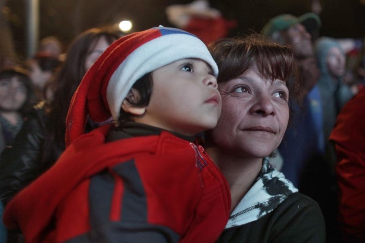 Una dona amb un nen a coll mira en una pantalla gegant de televisió l’inici de les operacions de rescat a la mina de San José.