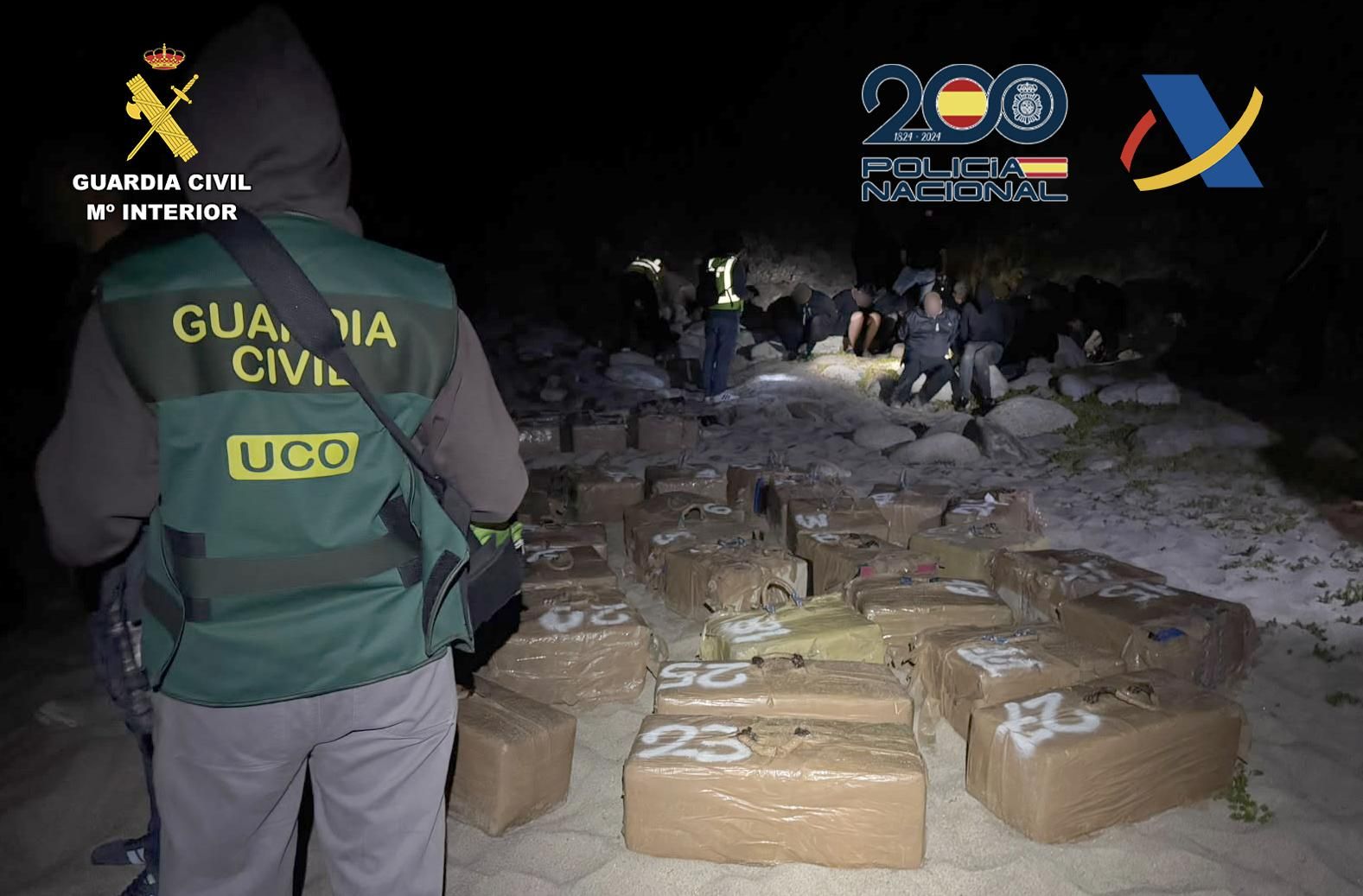 Un agente custodia parte del alijo intervenido en Muxía; al fondo, algunos de los detenidos en la operación.