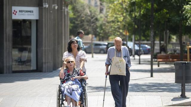 Una parella de grans passejant pel passeig San Juan, acompanyats d’una cuidadora que porta la cadira de rodes de la senyora. Barcelona, 29 de juliol del 2026. Foto: Belén González / EP