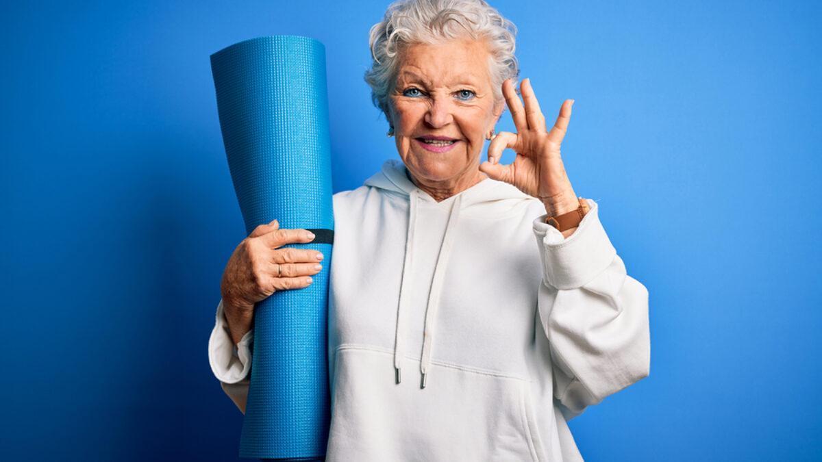 Senior beautiful sporty woman holding mat for yoga standing over isolated blue background doing ok sign with fingers, excellent symbol