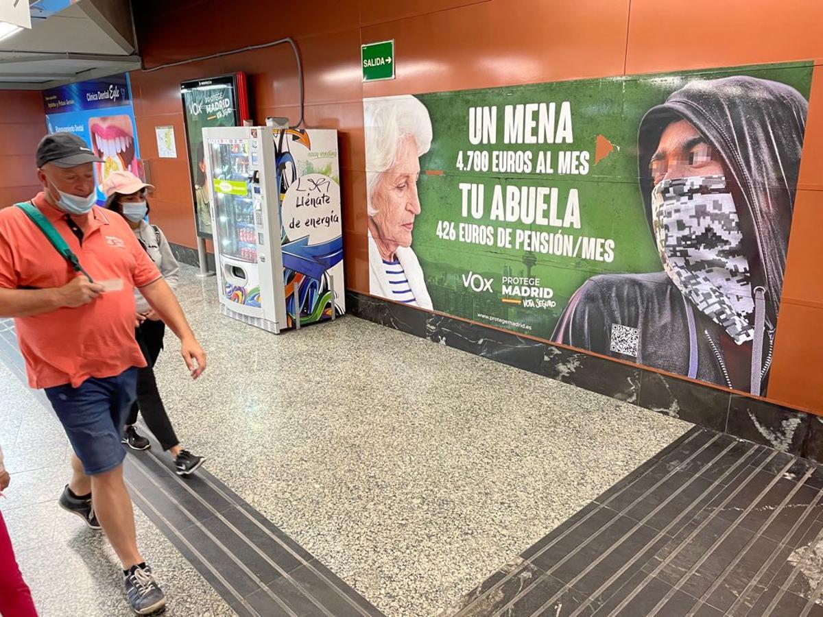 Usuarios del Metro y de Cercanías pasan junto al cartel electoral contra los menores no acompañados que ha instalado Vox en la estación madrileña de la Puerta del Sol.