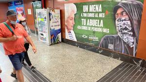 Usuarios del Metro y de Cercanías pasan junto al cartel electoral contra los menores no acompañados que ha instalado Vox en la estación madrileña de la Puerta del Sol.