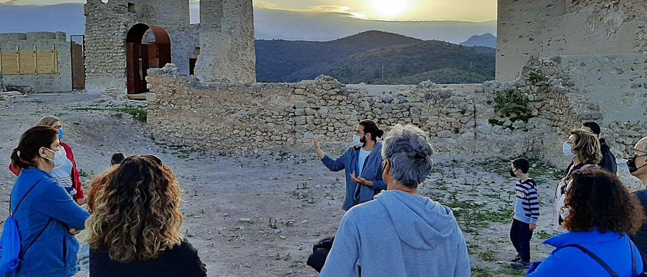 Un grupo de visitantes atiende las explicaciones del guía del Castell de Bairén en una puesta de sol. A la derecha, el acceso a la Cova del Parpalló, que aparece en la parte superior.  levante-emv