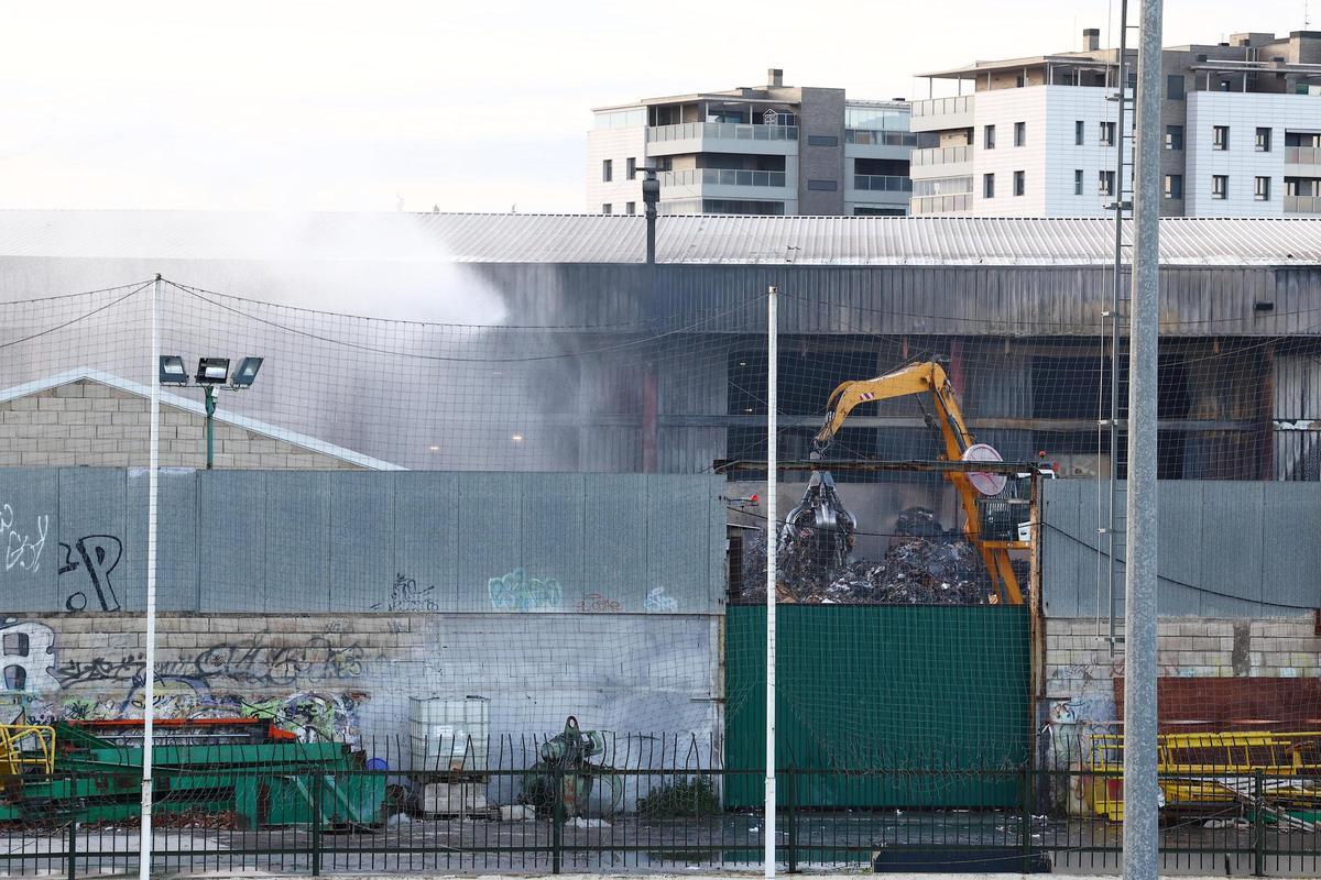 Una grúa trabaja tras el incendio en la planta de Saica en Zaragoza, con varios edificios residenciales de fondo.