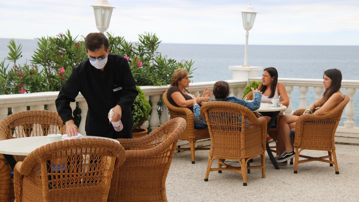 Un cambrer desinfectant les cadires de la terrassa de l&#039;Hotel Costa Brava amb unes dones prenent un cafè al darrera, en una imatge d&#039;arxiu