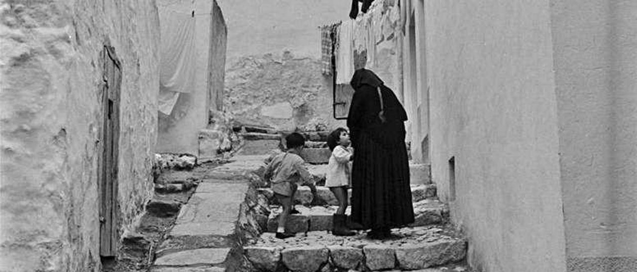 Una mujer, «vestida de negro y con pañuelo en la cabeza», junto a dos niños en «una callejuela estrecha, empedrada, escalonada y ascendente del barrio de sa Penya, con sus casas encaladas en blanco».