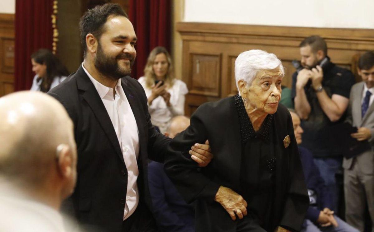 María del Carmen Canella recoge la medalla de oro a su abuelo del brazo de Miguel Ángel Bollo. Debajo, asistentes al acto, en el Paraninfo. | FERNANDO RODRÍGUEZ