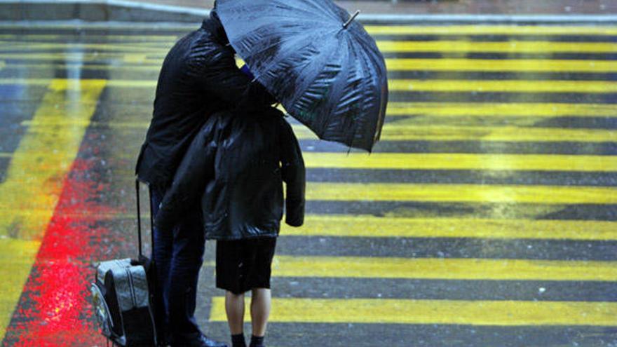 Un padre protege de la lluvia y el viento con un paragüas a su hijo en un paso de cebra.