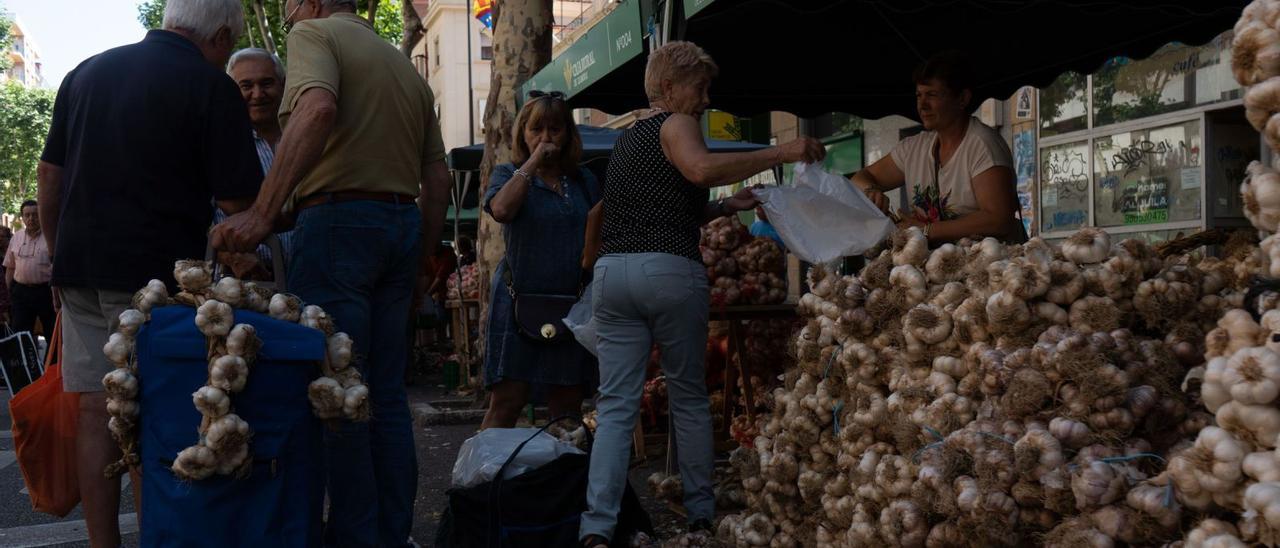 Un grupo de hombres carga con bolsas y carros de la compra con ristras de ajos mientras una vendedora ayuda a una clienta a hacer su compra. | Jose Luis Fernández