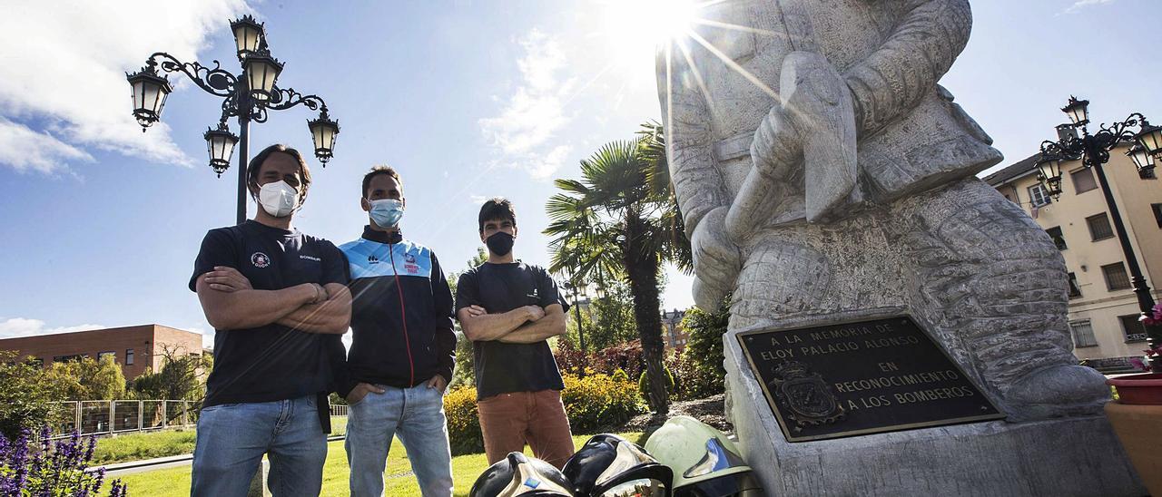 Por la izquierda, Aarón Igelmo, Emilio Martínez y Alberto Martínez, con sus cascos, junto a la estatua en recuerdo de Eloy Palacio, bombero fallecido en acto de servicio en Oviedo. | M. López