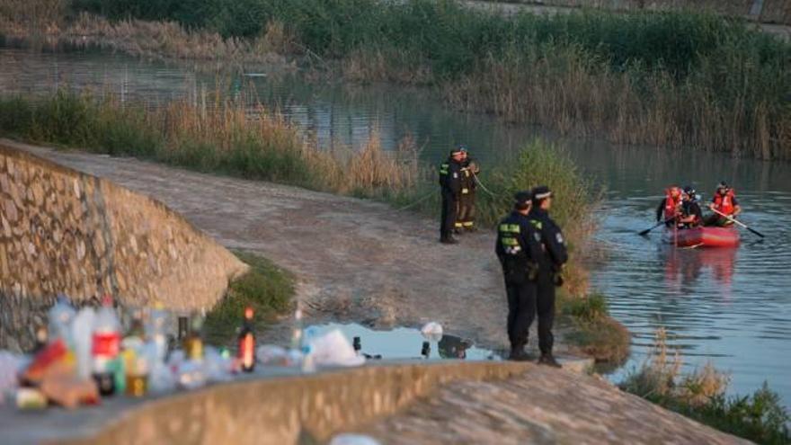 Dos policías locales observan a los Bomberos rastreando el cauce del río, con restos de &#039;botelleo&#039; en primer plano.