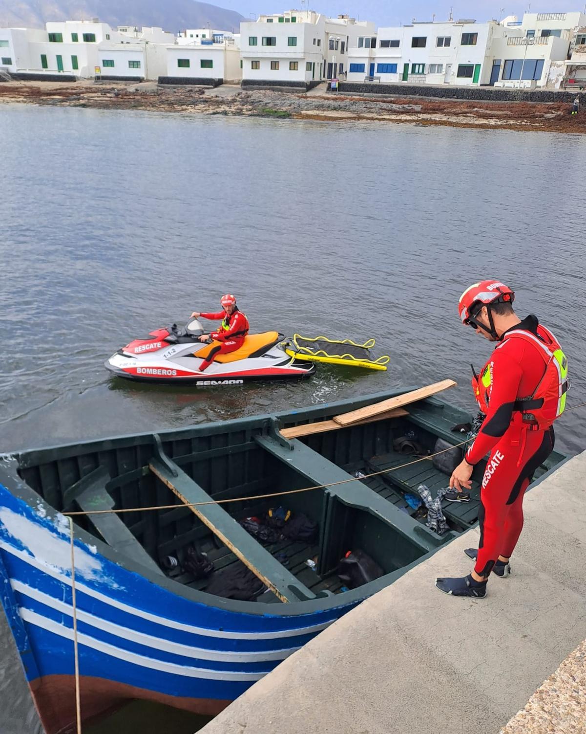 Dos agentes de bomberos junto a la patera atracada este jueves, 27 de abril, en el muelle de Famara.