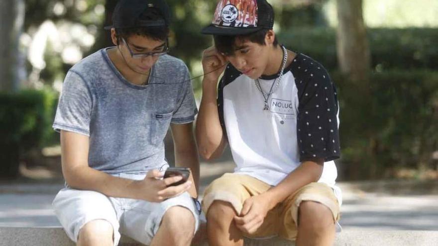 Dos jóvenes comparten música en su teléfono móvil sentados en un banco.