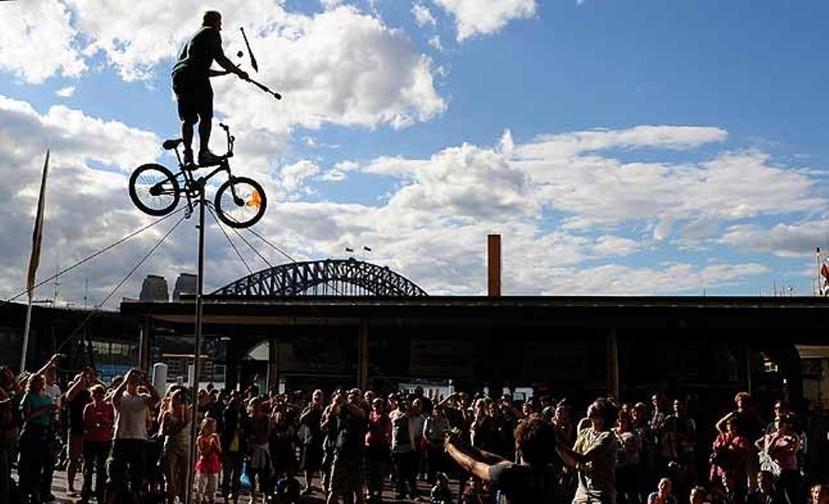 Un artista de teatre al carrer es balanceja en una bicicleta aèria sostinguda sobre un pal, mentre fa malabars amb una espasa, una torxa i una poma durant una actuació a Sydney, Austràlia.
