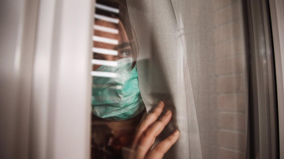 Una mujer con mascarilla observa tras una ventana.