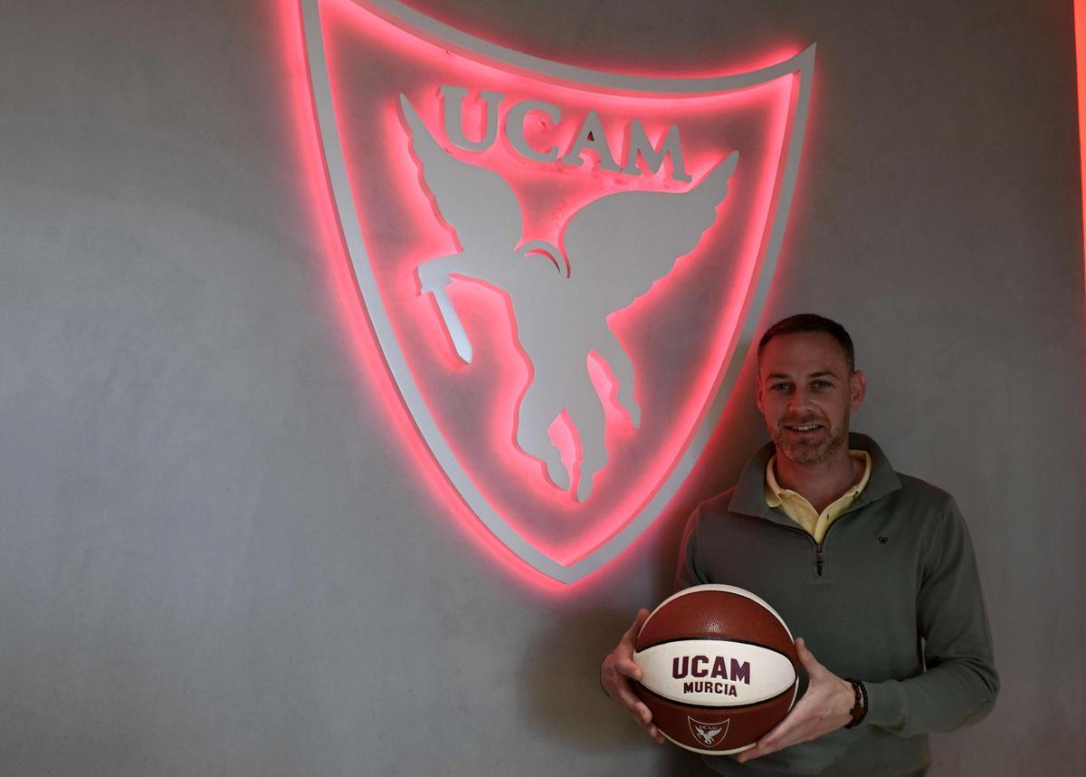 Miguel Ángel Noguera, concejal de Deportes del ayuntamiento de Murcia, posa junto a un escudo del UCAM Murcia CB