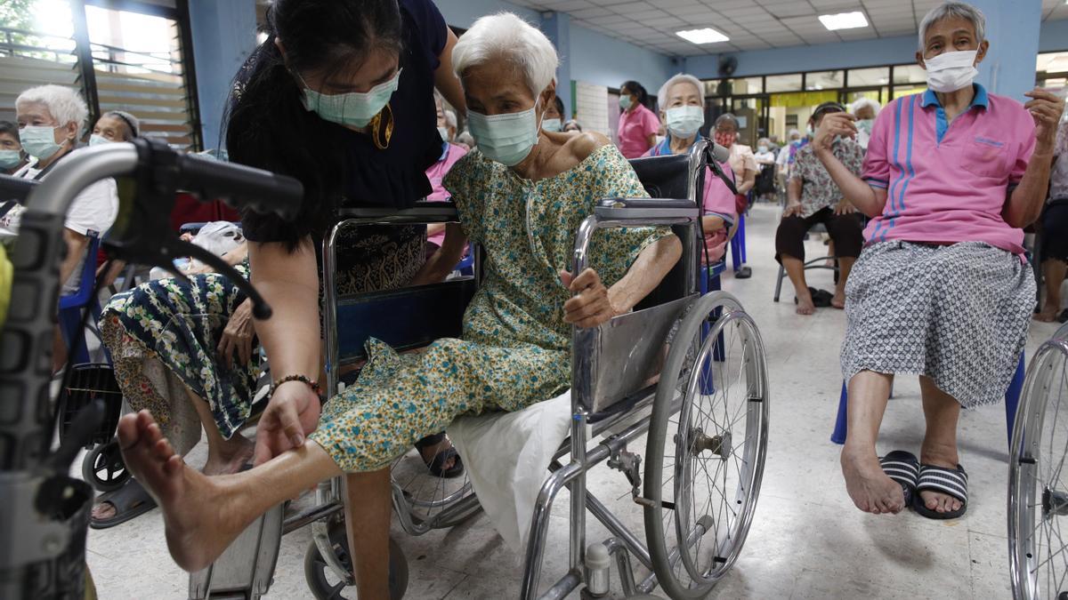 Una asistente ayuda a una anciana en silla de ruedas a levantar una pierna, durante los ejercicios matinales que realiza en la residencia Ban Bang Khae II en Bangkok.