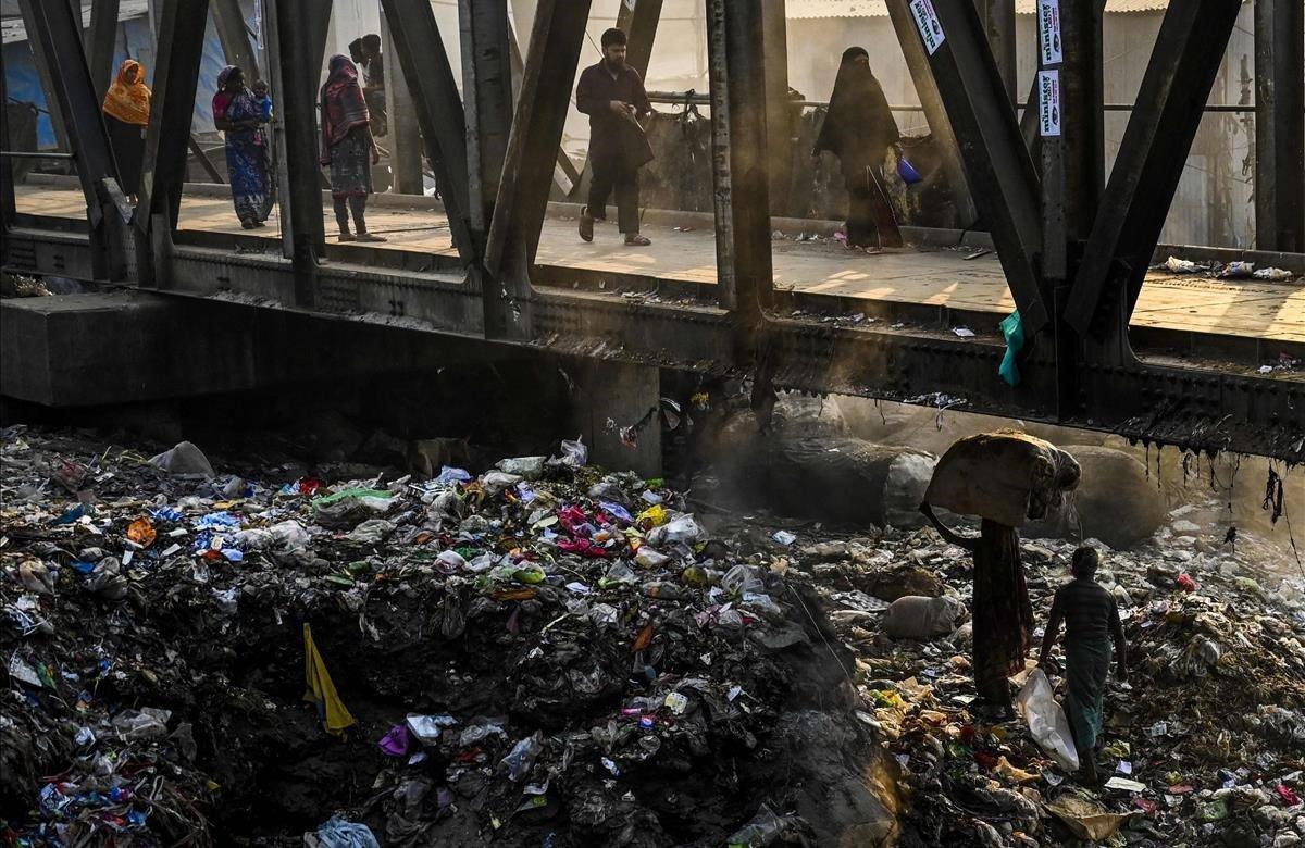 Una mujer de Bangladesh transporta desechos reciclables sobre pilas de basura debajo de un puente peatonal en Dhaka.