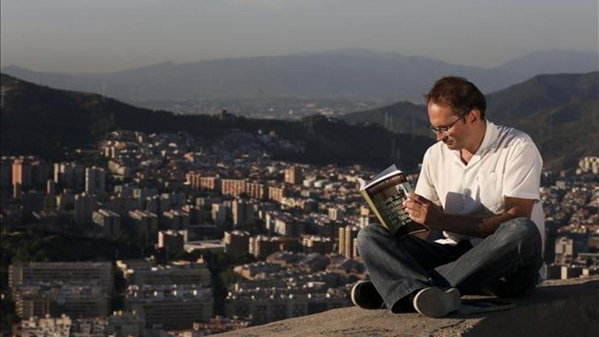 El líder de ICV, Joan Herrera, hojea 'Els barris de Barcelona', de Josep Maria Huertas Claveria, sentado en el mirador del Turó de la Rovira, con Torre Baró al fondo. JULIO CARBÓ