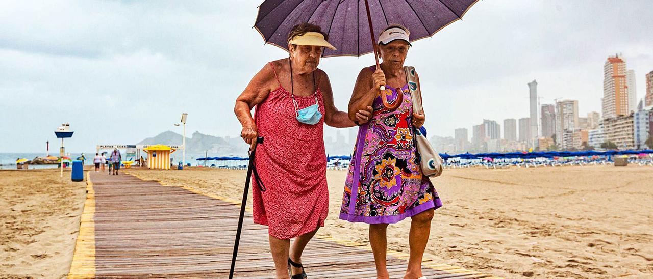 Dos señoras mayores salen ayer de la playa de Benidorm protegidas de la fina lluvia con un paraguas.