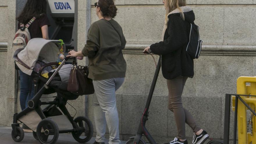 Una chica en patinete circula detrás de una mujer con un carrito de bebé.