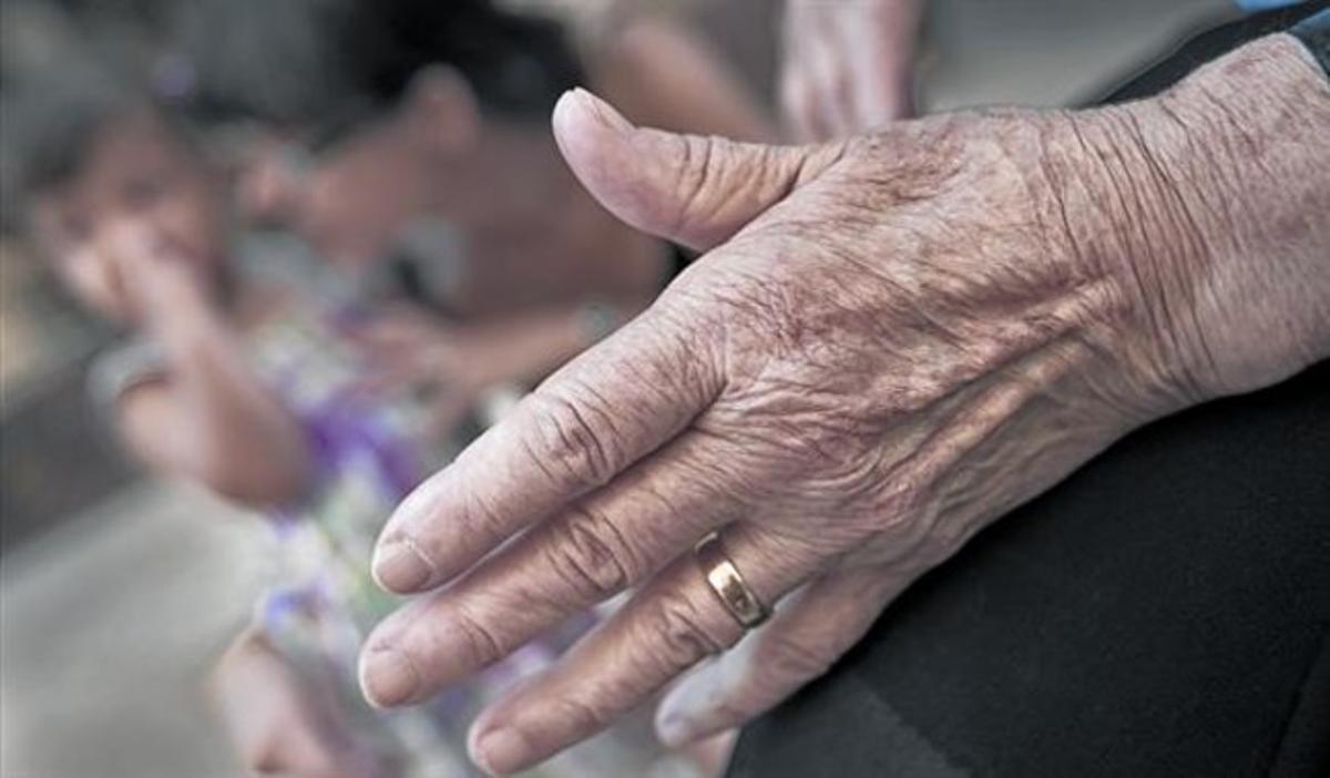 Primer plano de la mano de una anciana, con su hija y su nieta, sentadas al fondo, en Barcelona.